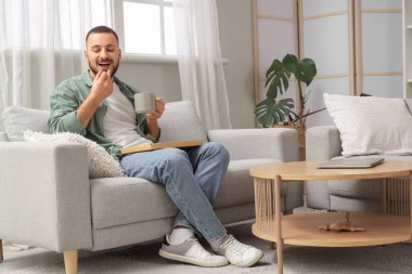 Evdeki kanepede bir fincan çayla çikolata yiyen genç adam.