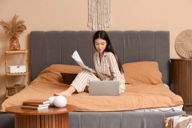 Pijamalı güzel bir kadın yatak odasında doküman ve dizüstü bilgisayarla çalışıyor.