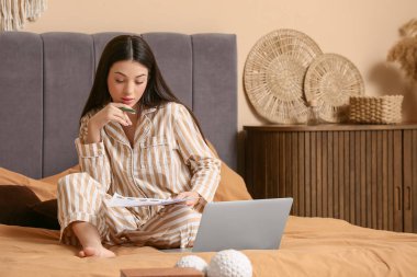 Pijamalı güzel bir kadın yatak odasında doküman ve dizüstü bilgisayarla çalışıyor.