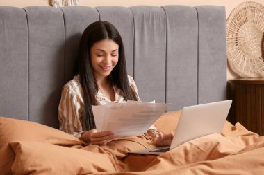 Pijamalı güzel bir kadın yatak odasında doküman ve dizüstü bilgisayarla çalışıyor.