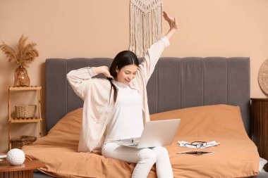 Dizüstü bilgisayarla çalışan ve yatak odasında oturan güzel genç bir kadın.