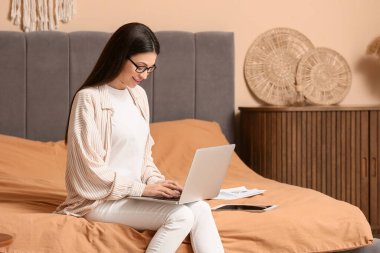 Dizüstü bilgisayarla çalışan ve yatak odasında oturan güzel genç bir kadın.
