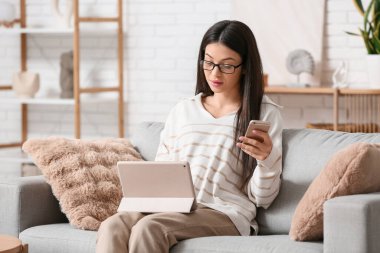 Modern tablet bilgisayarı ve cep telefonu olan güzel bir kadın oturma odasında koltukta oturuyor.