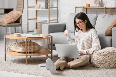 Oturma odasında dizüstü bilgisayar ve bir fincan kahveyle çalışan güzel genç bir kadın.