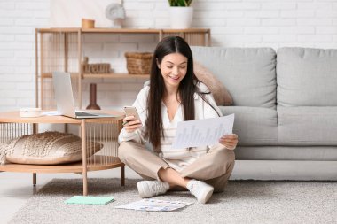 Oturma odasındaki halının üstünde dokümanlar ve cep telefonuyla çalışan güzel genç bir kadın.
