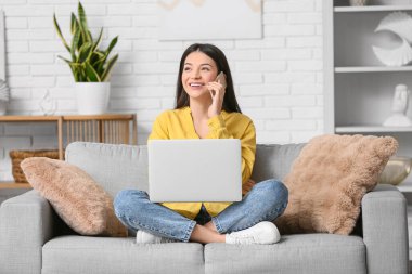 Güzel genç bir kadın cep telefonuyla dizüstü bilgisayarla oturma odasındaki kanepede oturuyor.