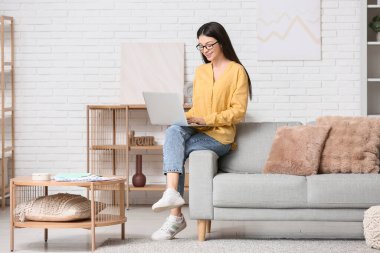 Dizüstü bilgisayarla çalışan ve oturma odasındaki kanepede oturan güzel genç bir kadın.