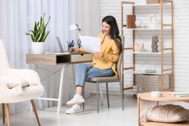 Oturma odasındaki masada doküman ve dizüstü bilgisayarla çalışan güzel genç bir kadın.