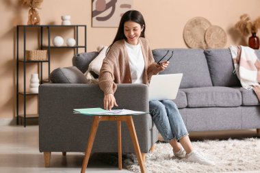 Oturma odasındaki kanepede doküman ve dizüstü bilgisayarla çalışan güzel genç bir kadın.