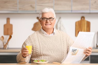 Yakışıklı son sınıf öğrencisi, mutfakta bir bardak meyve suyu ve taze salata ile gazete okuyor.