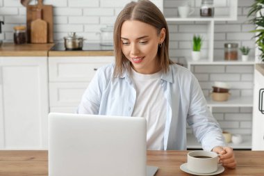 Mutfaktaki masada bir fincan sıcak kahve ve modern dizüstü bilgisayarı olan güzel bir kadın.
