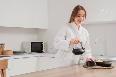 Bornozlu, çaydanlıklı, fincanlı ve kuru çay yapraklı genç kadın mutfakta.