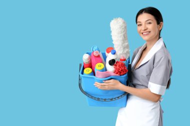 Güzel oda hizmetçisi elinde kovayla temizlik ürünleri ve mavi arka planda aletler tutuyor.