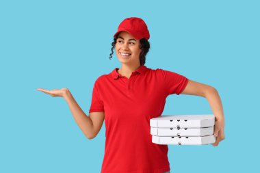 Afro-Amerikan kadın kurye elinde lezzetli pizzayla karton kutular tutuyor ve mavi arka plandaki bir şeyi işaret ediyor.