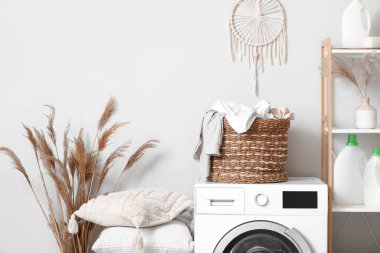 Modern çamaşır makinesi, raf ünitesi ve çamaşır sepeti banyoda beyaz duvarın yanında.