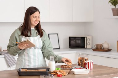 Mutfaktaki masada not defteri ve kurabiyeleri olan genç bir kadın.