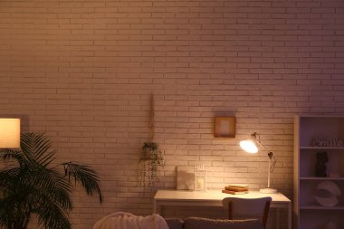 Masası, sandalyesi ve beyaz tuğla duvarın yanında parlayan lambası olan karanlık oturma odasının içinde.