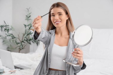 Evdeki yatakta aynaya bakarken maskara süren genç ve güzel bir kadın.