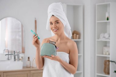 Havluya sarılı genç bir kadın banyoda makyaj çantasından rimel alıyor.