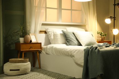 Komodinin yanında saksı, çerçeve, kanepe ve gece yatağının yanında lamba var.