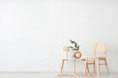 Kahve masalı rahat ahşap sandalye, okaliptüs dalları vazosu ve beyaz tuğla duvarın yanında lamba.