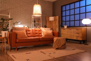 Modern oturma odasının içinde kahverengi kanepe, çekmeceler, raf üniteleri ve akşamları parlayan lambalar