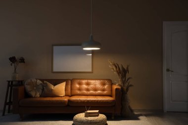 Karanlık oturma odasının içinde kahverengi kanepe, boş çerçeve ve geceleri parlayan lamba.
