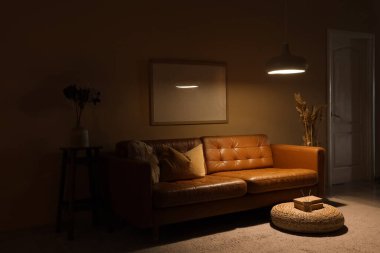 Karanlık oturma odasının içinde kahverengi kanepe, boş çerçeve ve geceleri parlayan lamba.