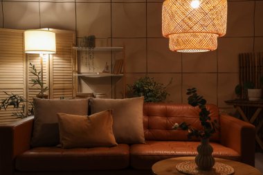 Kahverengi kanepesi, bitkileri ve geceleri parlayan lambaları olan karanlık bir oturma odası.