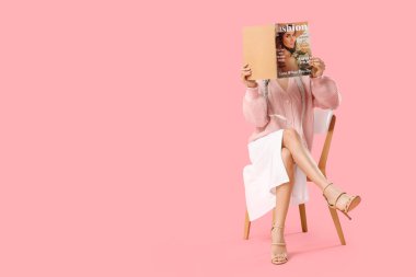 Güzel, genç ve şık bir kadın pembe arka planda otururken dergi okuyor.