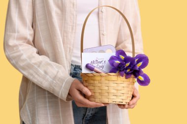 Sarı arkaplanda anneler günü için çiçek, kartpostal ve kozmetik taşıyan sepet taşıyan genç bir kadın.