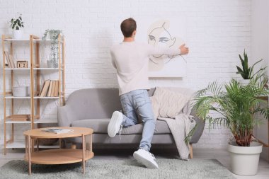 Genç adam oturma odasındaki beyaz tuğla duvara resim asıyor.