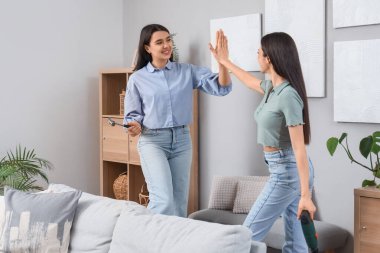 Aletleri olan genç kadınlar evlerinde duvarlara asılı duran tabloların yanında birbirlerine beşlik çakıyorlar.