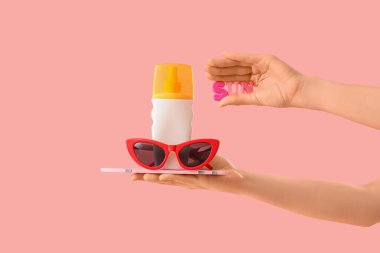 Güneş gözlüklü ve pembe arka planda güneş kremi şişesi tutan kadın.