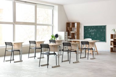 Sınıfın içinde karatahta, raf ünitesi ve masalar var. Okul yılı sonu.