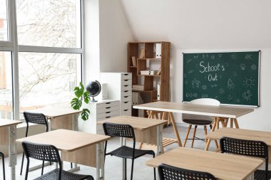 Sınıfın içinde karatahta, raf ünitesi ve masalar var. Okul yılı sonu.