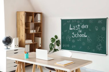 Sınıfın içinde karatahta, raf ünitesi ve öğretmen masası var. Okul yılı sonu.