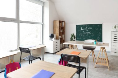 Sınıfın içinde karatahta, raf ünitesi ve masalar var. Okul yılı sonu.