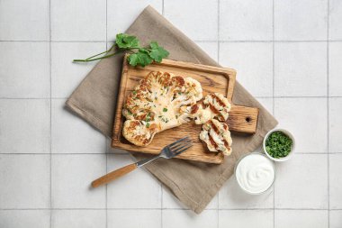 Tahta tahtada maydanozlu ızgara karnabahar bifteği ve beyaz fayanslı bir kase sos.