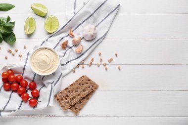 Bir kase lezzetli humus, beyaz ahşap arka planda malzemeler, krakerler ve peçete.