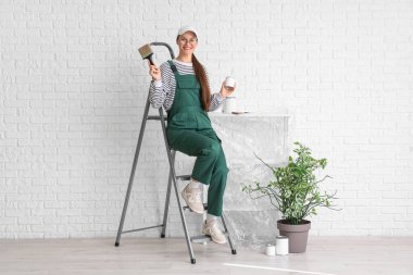 Boya fırçası ve beyaz tuğla duvarın yanındaki merdivende oturan kadın işçi.