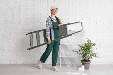 Beyaz tuğla duvarın yanında merdiven taşıyan bir kadın işçi.
