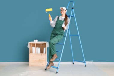 Mavi duvarın yanında boya silindirli ve merdiveni olan kadın işçi.