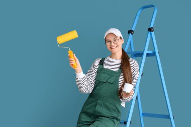 Mavi duvarın yanında boya silindirli ve merdiveni olan kadın işçi.