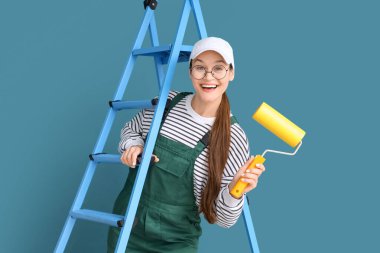 Mavi duvarın yanında boya silindirli ve merdiveni olan kadın işçi.