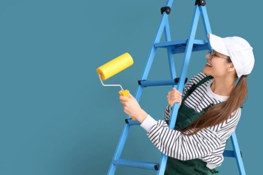 Mavi duvarın yanında boya silindirli ve merdiveni olan kadın işçi.
