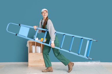 Mavi duvarın yanında merdiven taşıyan bir kadın işçi.