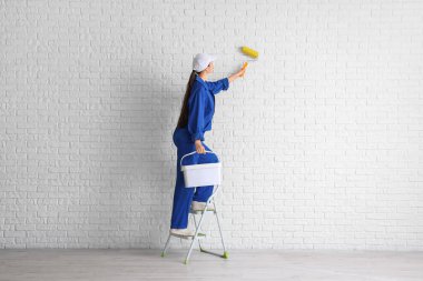 Beyaz tuğla duvarın yanındaki merdiven üzerinde boya silindiri ve kova taşıyan kadın işçi.