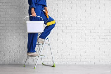 Beyaz tuğla duvarın yanındaki merdiven üzerinde boya silindiri ve kova taşıyan kadın işçi.