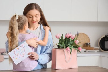 Sevimli küçük bir kız annesine sarılıyor ve anneler gününü lalelerle ve mutfakta kartpostalla kutluyor.